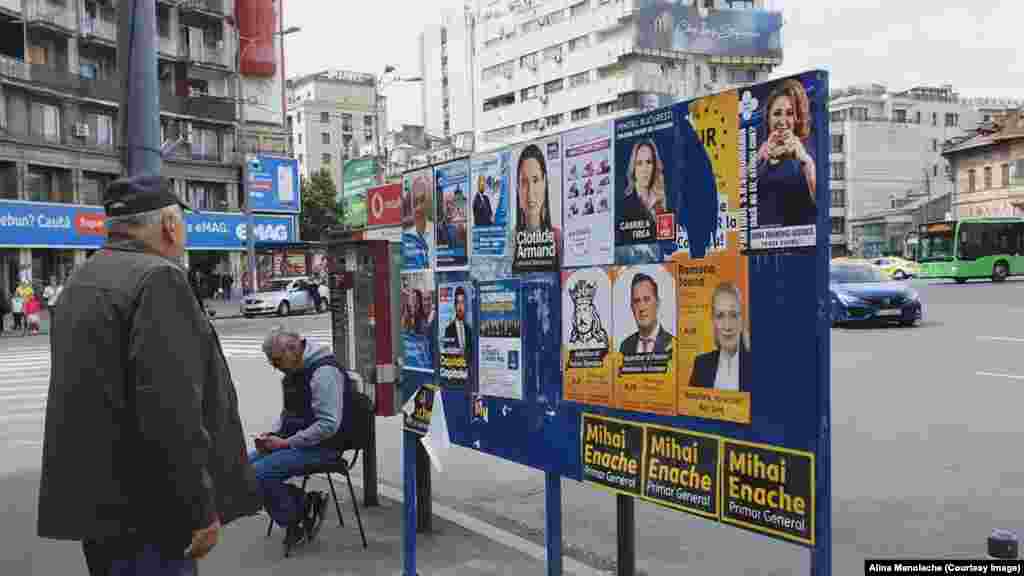 Panou cu afișe electorale în Piața Romană. Un bărbat privește promisiunile electorale care răzbat din mesajele candidaților. Lângă panou, proprietarul florăriei de vizavi stă pe un scaun, așteptând clienții. 