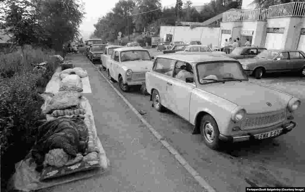 Refugiați est-germani dorm pe trotuar lângă mașinile lor Trabant în Budapesta. Odată cu schimbarea spectaculoasă a Ungariei în direcția libertății, în 1989, mii de est-germani care suportau supravegherea constantă a poliției secrete Stasi au început să plece spre est. Ungaria era unul din „statele socialiste frățești” în care est-germanii aveau voie să călătorească.