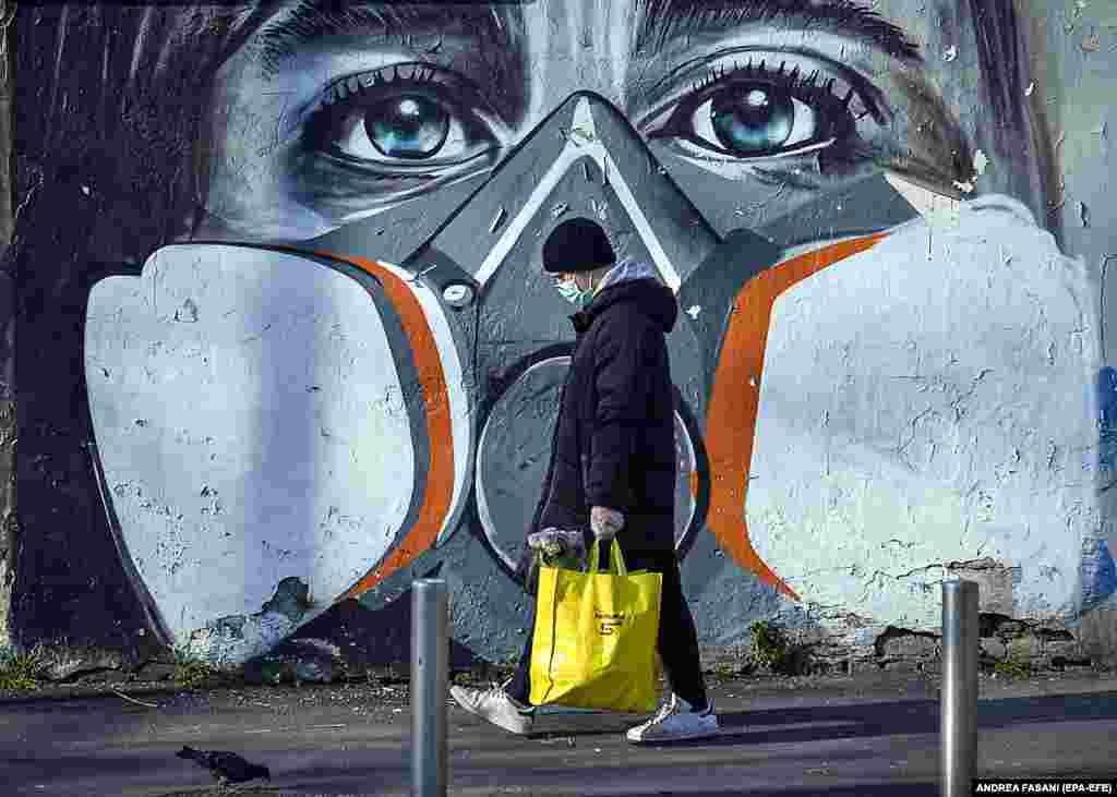 Një burrë italian me një maskë në fytyrën e tij kalon pranë një pikture murale në Milano, që portretizon po ashtu një person me maskë.